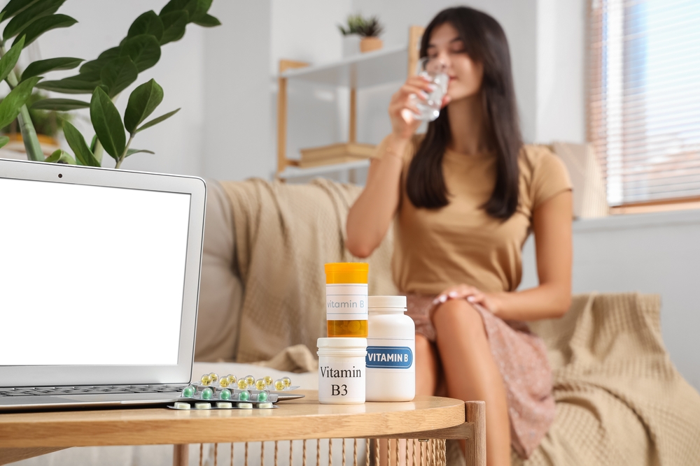 Young woman taking pills at home, focus on bottles with different vitamins B and laptop