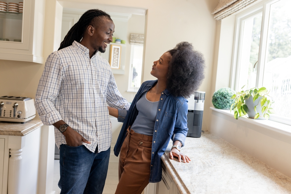 African American couple smiling and talking in kitchen near sunny window. Family, lifestyle, happiness, conversation, breakfast, home
