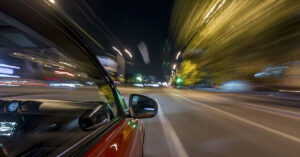 Most Drivers Miss This Simple Button That Improves Visibility At Night car driving at high speed on a city street at night. The motion blur suggests a long-exposure or a hyperlapse effect, giving a sense of rapid movement. The city lights and streetlights create a blur of colors, reflecting off the car's body and the wet road.