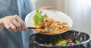 How France Is Fighting Food Waste Through Supermarket Donations person holding a plate with leftover food—some pasta with sauce and a piece of lettuce—over a trash bin. The person is using a fork to scrape the food off the plate into a black trash bag inside the bin. The background is softly lit with natural light coming through a window or curtain. The overall scene suggests discarding leftover food waste.