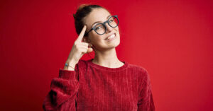 8 Traits That Indicate a High IQ, Even if You Don’t Realize It red background with woman wearing a red sweater with right index finger pointed to her forehead. Smart/intelligence concept