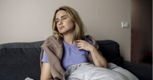 COVID’s Long Shadow: Signs the Virus May Still Affect You Years Later A woman with blonde hair wearing a purple t-shirt sits wrapped in a brown sweatshirt and blanket on a dark gray couch, looking off into the distance with a somber or unwell expression.