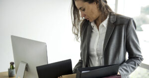 Companies Say AI Is Behind Layoffs, But the Story Isn’t That Simple A woman in business attire places folders and office supplies into a cardboard box on her desk, as if packing up her workspace. A computer monitor is on the desk beside her.
