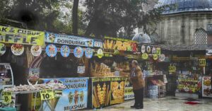 Family Dies After Suspected Food-Related Illness During Holiday in Turkey A street vendor stall in a busy outdoor market selling various snacks and drinks, with colorful signs in Turkish advertising items like dondurma (ice cream), waffles, milkshakes, and tea. A man in a brown jacket is selecting food from the display.