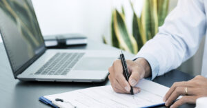 Person writing on a clipboard with a pen at a desk, with an open laptop in the background.