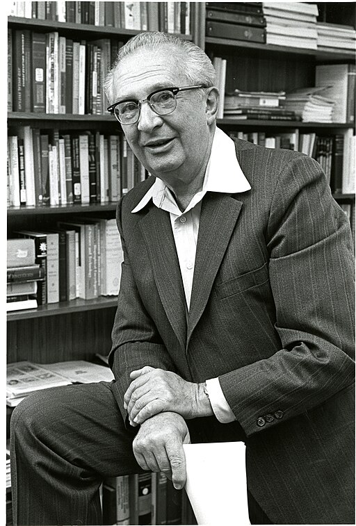 Jacob Rabinov sits in his office before a wall of bookshelves in this 1950s black and white photograph. The inventor wears glasses and a dark suit with a light colored shirt. His photocell device automatically adjusted car mirrors to reduce headlight glare at night.