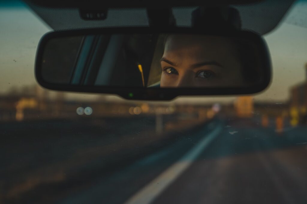 A driver's eyes reflected in a car's rearview mirror at dusk, with blurred city lights visible through the windshield. The mirror displays a small green indicator light for its auto-dimming feature.