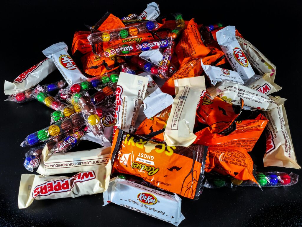 Assorted Halloween candy spread on a black surface. Includes Reese's, Kit Kat, Smarties, and various chocolate bars in orange, white, and colorful wrappers.