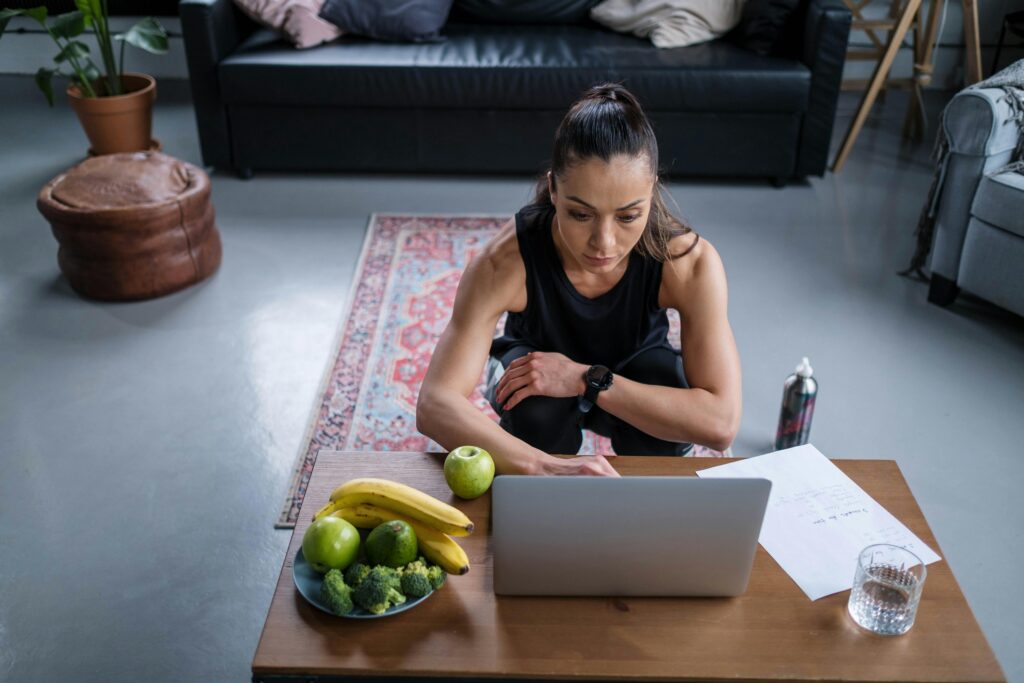 A woman in workout clothes sits on the floor using a laptop with fruit, broccoli, and a glass of water on the table.