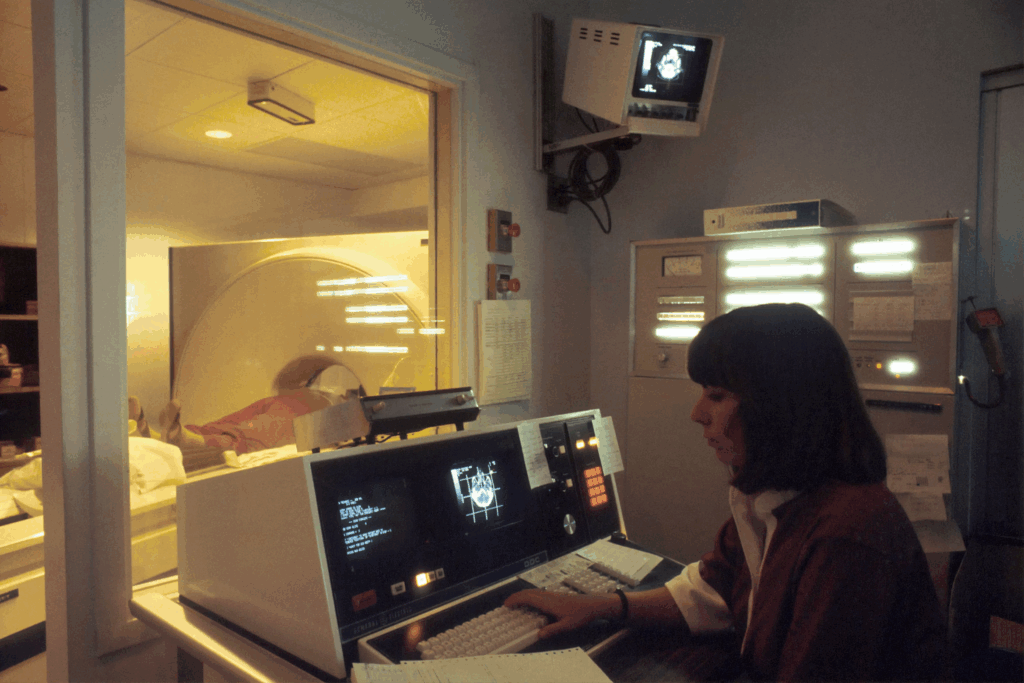 A computer-assisted tomographic (CAT) scanner, with a Caucasian female technician working at a screen and behind a glass wall. A patient is on a table and being tested by the CAT scanner. The lighting is very subdued.