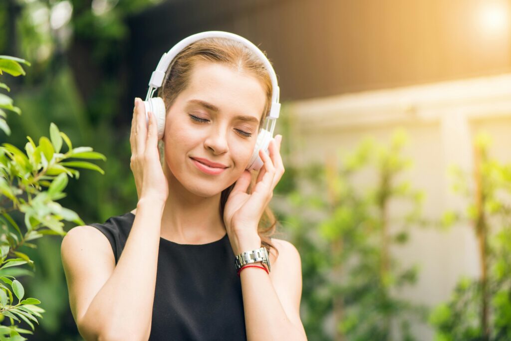 A woman stands outdoors among green plants, eyes closed and hands resting on her white headphones, smiling softly.
