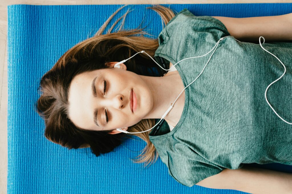 A woman lies on a blue yoga mat with her eyes closed and white earbuds in, looking relaxed and content.