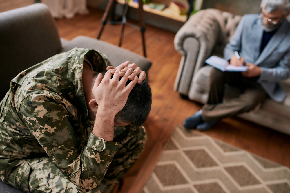 Middle aged military man holding his head in pain and depression during therapy session with psychologist. Soldier suffering from psychological trauma. PTSD concept. Focus on military. High angle