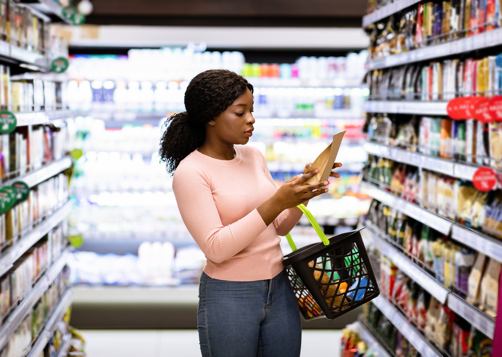 Beautiful African American woman making choice of products at supermarket. Focused female consumer shopping for groceries, selecting food, reading labels at modern mall
