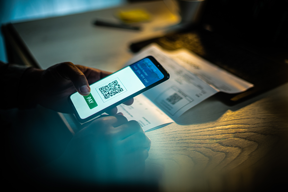Close up of an African American male hand holding mobile phone with QR payment app on screen, at a desk with a laptop and a bill