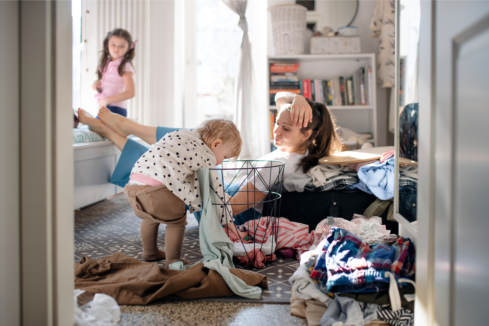 Tired mother with small toddler daughter in messy bedroom at home, resting.
