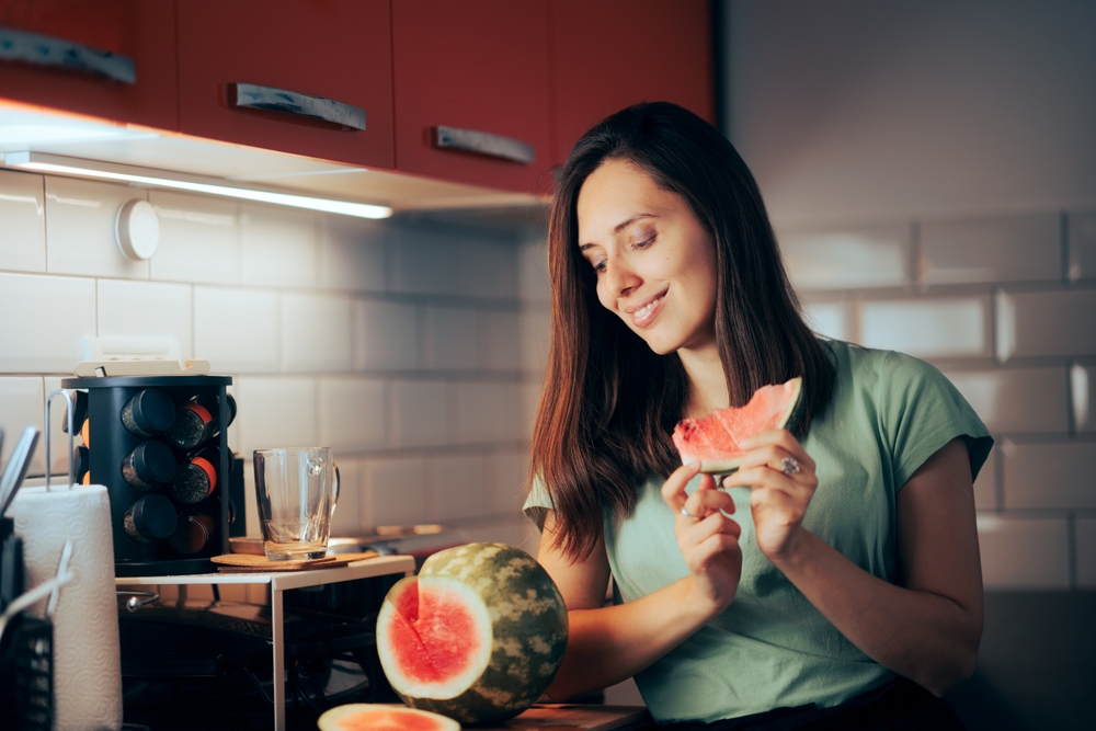 
Woman Eating a Fresh Watermelon in the Evening. Hungry lady having a fruit snack in her kitchen 
