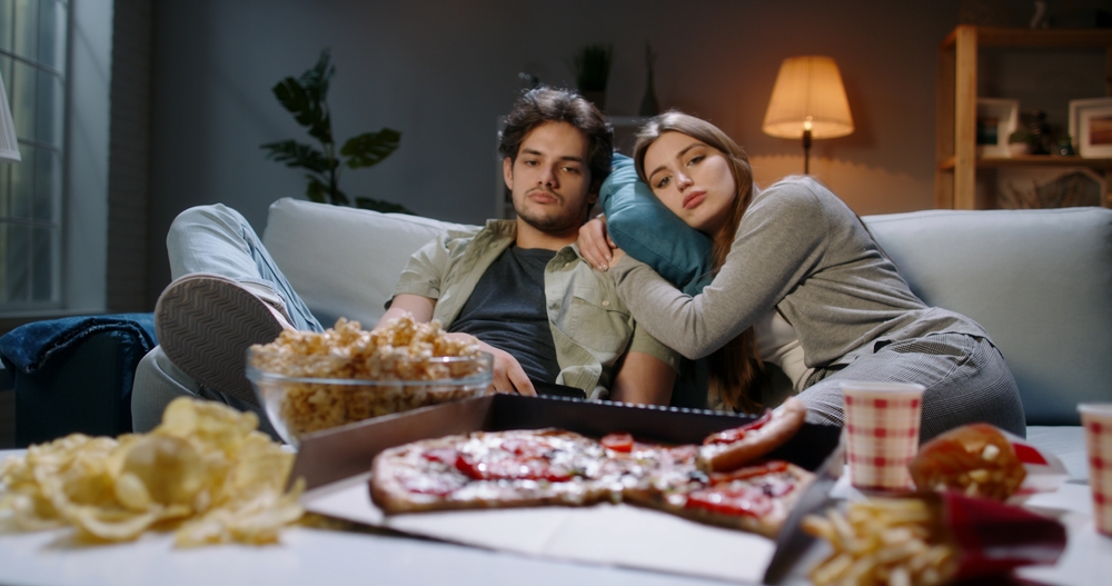 A young Caucasian couple was very upset. Bored students sitting in front of the TV, changing channels on the remote and eating unhealthy food, tired after a hard day