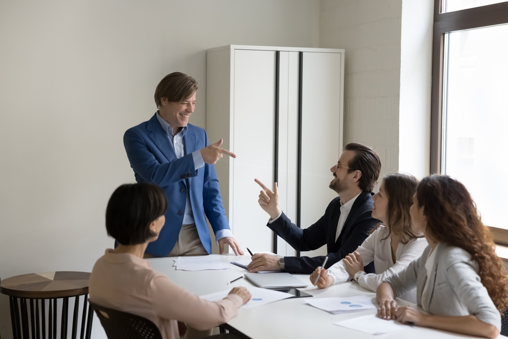 Happy mature boss in suit points finger to colleague, makes exactly gesture, agreed with subordinate opinion feels satisfied with briefing in modern conference room. Successful meeting, companionship
