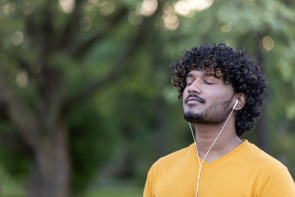 Close-up photo of a young Indian male athlete wearing headphones standing in a park listening to music and resting with his eyes closed.