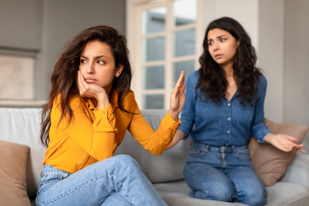 Annoyed lady making stop gesture to her friend talking after quarrel, refusing to do favors, setting healthy boundaries in friendship, sitting on sofa in modern living room indoor