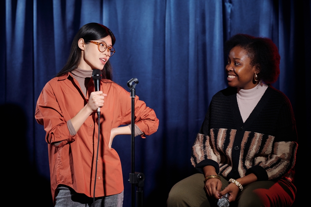 An Asian woman speaking into a microphone and looking at an African American artist sitting against the blue curtains of the stage