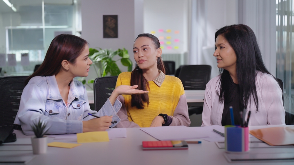 Indian smiling young adult woman business talking break time together indoor workplace. Teen age female corporate sitting office chair colleague speak group nodding listening gossip modern company 