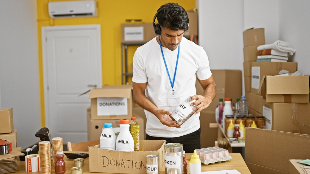 A focused hispanic man organizes canned beef donations in an indoor warehouse setting, exuding a helpful and professional vibe.