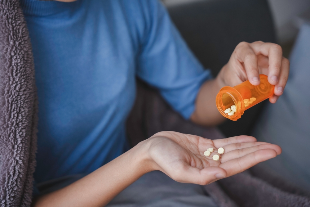 A sick woman is sitting on a bed under a blanket and pouring pills into a doctor's bottle with her hand