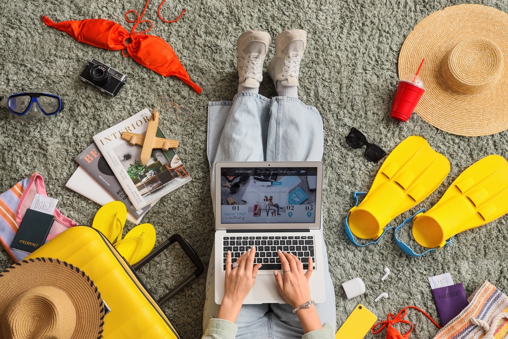Female travel blogger using laptop with beach accessories on green carpet, top view