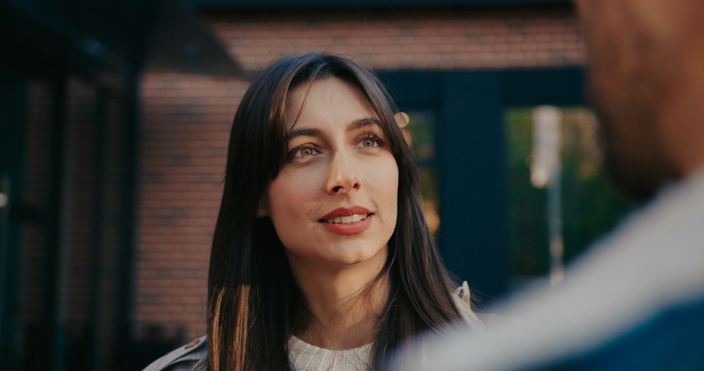 Portrait of pretty Caucasian woman talking with someone at street. Sunlight shining bright directly on face. People actively communicating with each other. During conversation girl nodding her head.