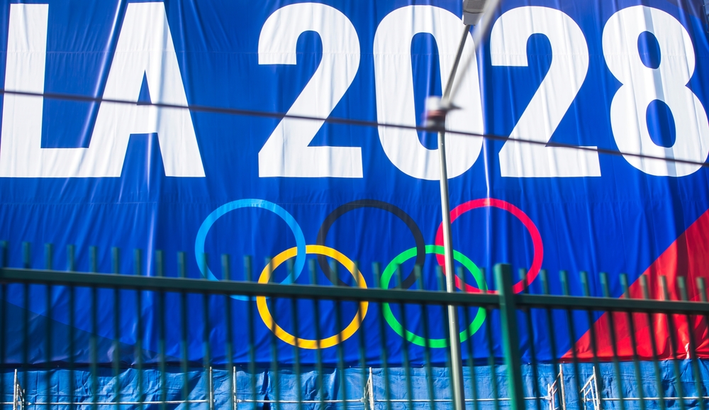 2024-11-20 Los Angeles USA
A promotional banner for the LA 2028 Olympics featuring the Olympic rings, displayed on a blue background with a green fence below.