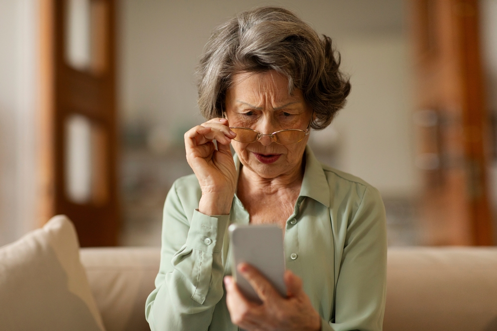 Bad vision health problems. Senior woman squinting eyes while looking at phone screen, touching eyeglasses, sitting on couch at home