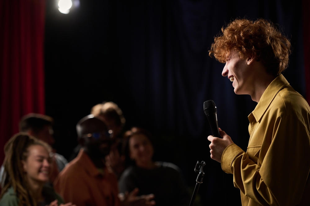 A person with curly hair holding a microphone while doing stand-up comedy on stage. A diverse audience enjoys the show and laughs together in a dark room