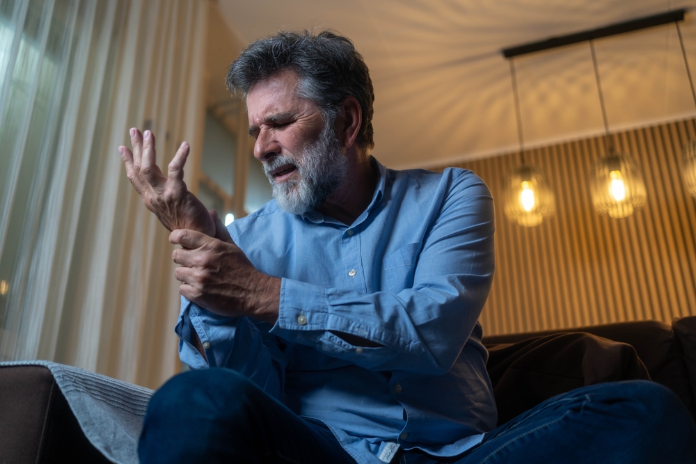 Elderly man with arthritis rubbing painful hands at home. Senior suffering from joint pain, rheumatism or stiffness, healthcare and aging concept.