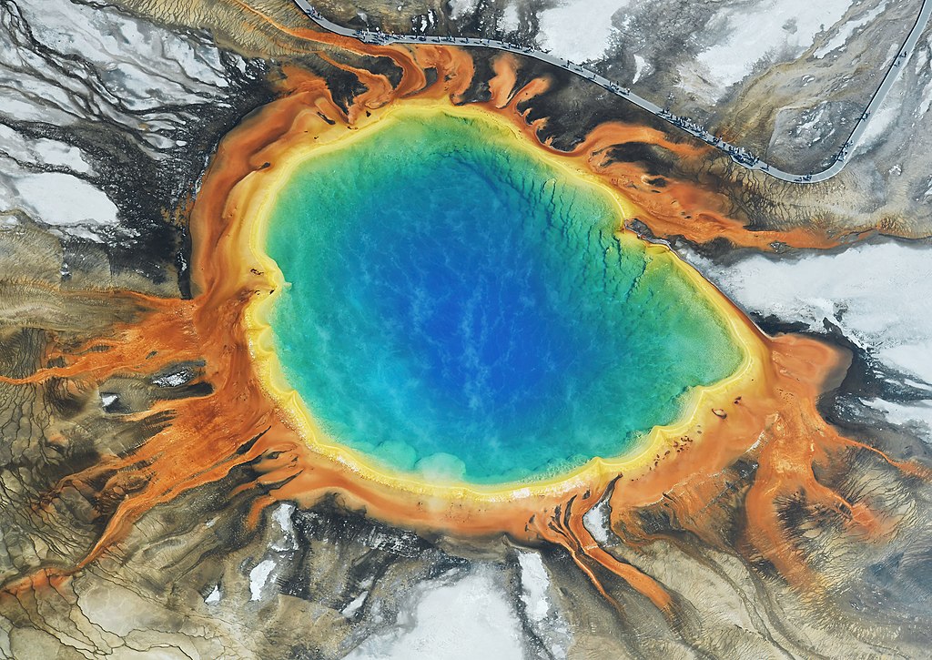 Aerial photograph of Grand Prismatic Spring showing its brilliant blue center surrounded by concentric rings of orange, yellow, and green created by heat-loving microorganisms, with white mineral deposits and snow visible at the edges.