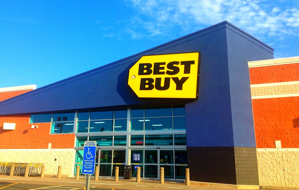 Best Buy storefront with the yellow price tag logo against a blue building, clear sky overhead.