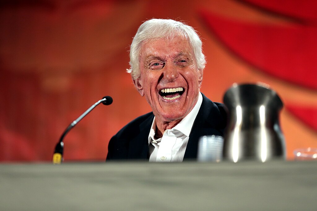 Dick Van Dyke laughing at a panel event, his white hair swept back and his face lit with joy.