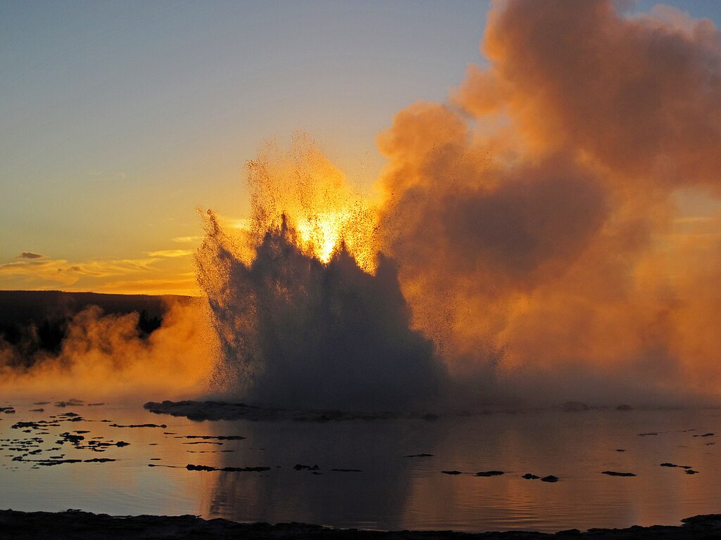 Silhouette of Great Fountain Geyser erupting against an orange and gold sunset sky, with water and steam exploding upward and reflecting on the surrounding pool.