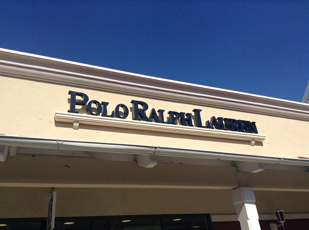 Polo Ralph Lauren store sign in dark lettering against a beige stucco wall and blue sky.