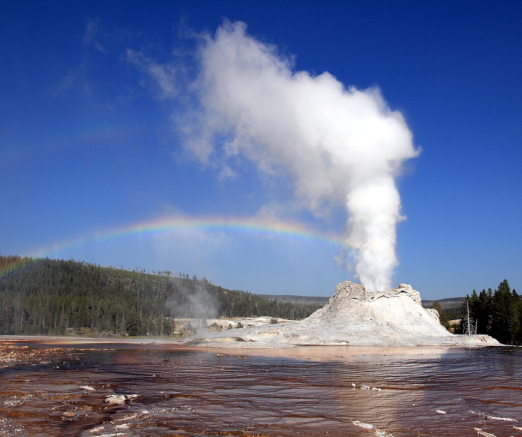 Photograph of Castle Geyser erupting with a tall column of white steam rising from its craggy cone, a rainbow arcing across the bright blue sky to the left, and rust-colored mineral deposits and shallow water in the foreground.