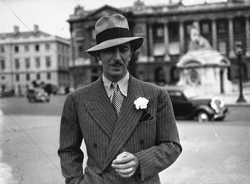 Walt Disney in a pinstripe double-breasted suit and fedora, with a white boutonniere on his lapel, photographed in black and white at a European plaza.