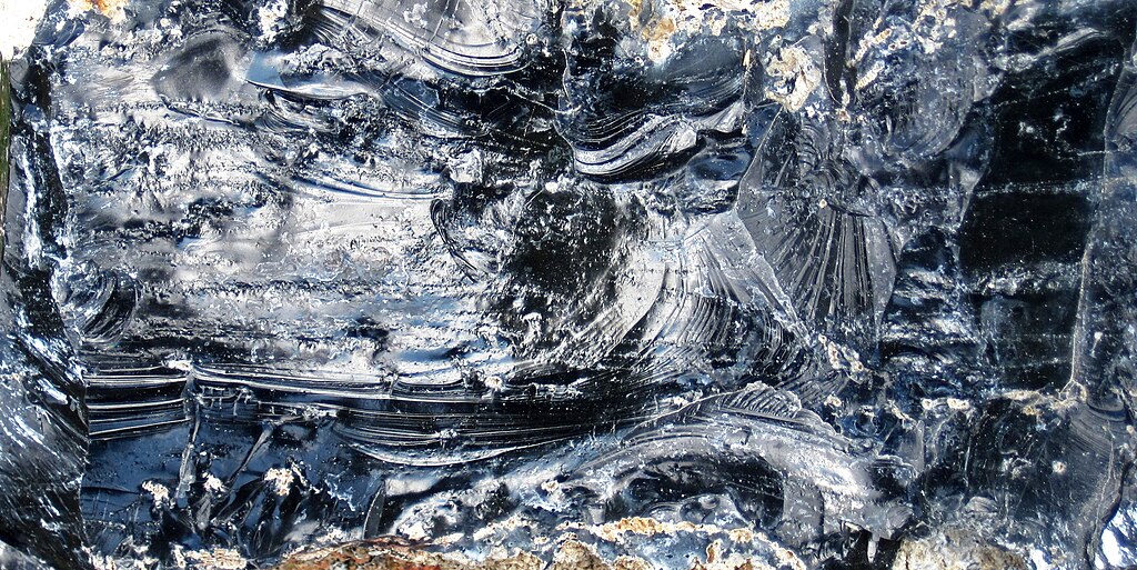 Close-up photograph of black obsidian volcanic glass with shiny, reflective surfaces showing conchoidal fracture patterns and some lighter gray inclusions.