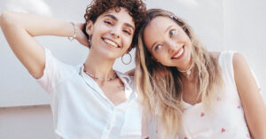 What Is Berrisexuality? Inside the Meme That Turned Into an Identity Two young women smiling broadly at the camera outdoors in bright sunlight. The woman on the left has short, curly brown hair and wears a white top and beaded necklaces, while the woman on the right has long blonde hair and wears a white dress with small pink heart patterns.