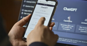 A person holds a smartphone displaying the ChatGPT interface, with a finger pointing at the screen. In the background, a computer monitor shows a larger ChatGPT homepage with sections such as “Capabilities” and “Limitations.”