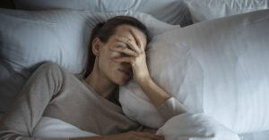 woman lying in bed with her eyes closed, appearing to be in discomfort or distress. She has one hand covering part of her face, suggesting she might be experiencing a headache, stress, or fatigue. The setting is a bed with white pillows and white bedding, and the lighting is soft and subdued, giving the scene a calm but somber mood. The woman's expression and body language convey a sense of tiredness or emotional strain.