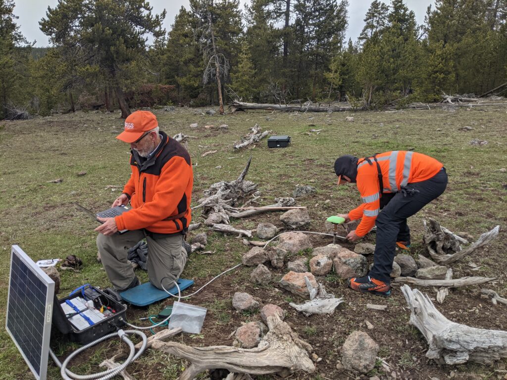 Two researchers in orange USGS safety gear working in a grassy clearing, one kneeling with a laptop connected to equipment, the other bending over rocks, with a solar panel and dead wood visible nearby.