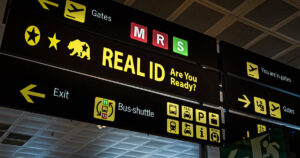 TSA to Charge $45 for Travelers Without a REAL ID Airport-style signage displaying various yellow directional icons for gates, exits, bus shuttles, taxis, parking, and metro. In the center, a prominent sign reads ‘REAL ID – Are You Ready?’ with a star and bear symbol above it.