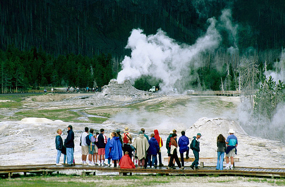 Photograph of a group of tourists standing on a boardwalk viewing area, backs to the camera, watching a geyser erupting in the middle distance with steam rising against a backdrop of dead trees and forest.