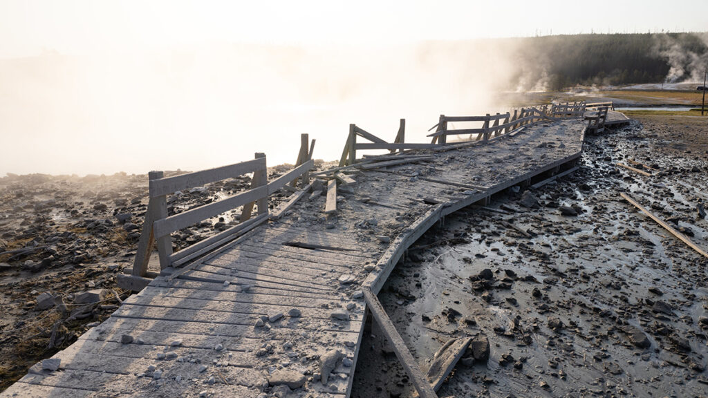Photograph of a wooden boardwalk covered in gray mud and debris, with warped and displaced planks, steam rising in the background from thermal features in a barren landscape.